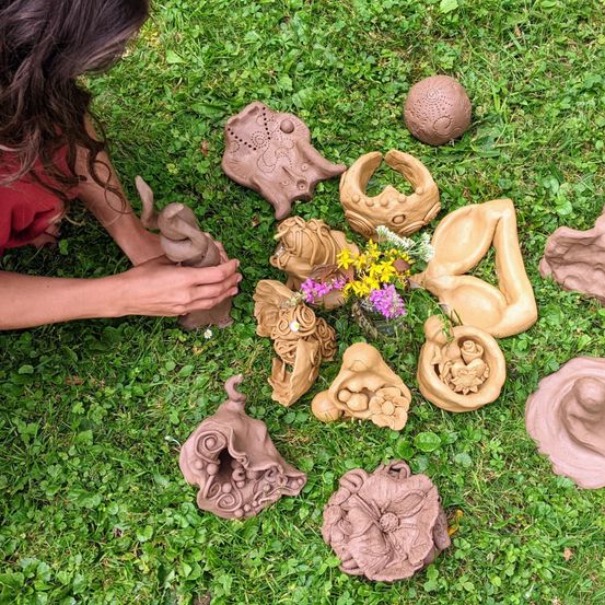 Eine Person kniet auf Gras und ordnet verschiedene Tonfiguren und Skulpturen an, einige stellen menschliche Formen und Tiere dar, mit Blumen in der Mitte.