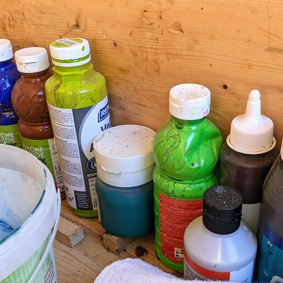 Verschiedene Flaschen mit Farbe, darunter grün, braun, blau und weiß, sind auf einem Holzregal aufgereiht. Ein weißer Eimer, gefüllt mit Farbe, steht links und ein weißes Tuch ist rechts platziert.