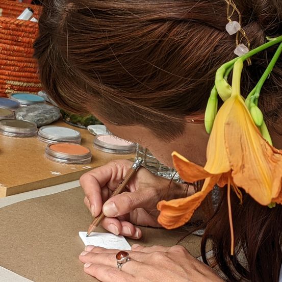 Eine Frau malt mit einem Bleistift auf einem Blatt Papier, während sie eine Blumenhaarspange trägt. Verschiedene Farben und Steine liegen auf dem Tisch.