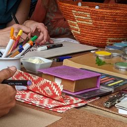 Ein Arbeitsbereich mit einer Vielzahl von Künstlerutensilien, darunter ein großer Korb, eine Sammlung von Malpinseln und eine Palette mit zahlreichen kleinen Farbtöpfchen. Ein lila Buch und eine Rolle Geschenkpapier sind ebenfalls sichtbar.