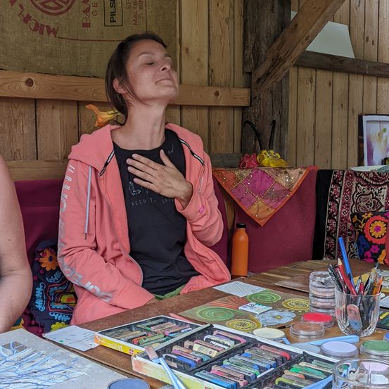 Eine Frau sitzt in einem Holzraum, die Augen geschlossen und die Hände auf der Brust. Verschiedene Kunstmaterialien liegen auf einem Tisch vor ihr. Eine weitere Person ist teilweise auf der linken Seite sichtbar.