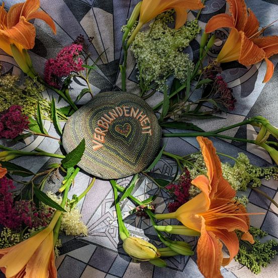 Ein Mosaikstein mit der Aufschrift 'NIE VERBUNDEN' ist von orangefarbenen Lilien und anderen Blumen auf einer gefliesten Oberfläche umgeben.