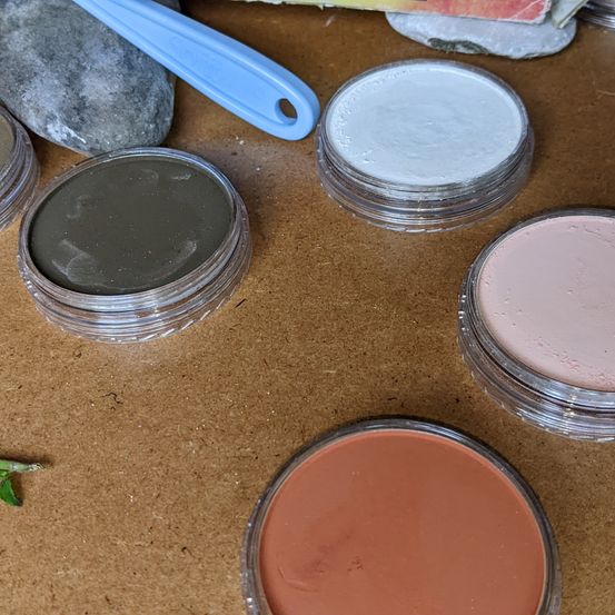 Verschiedene Farben von Mineralfarbe in Glastöpfen auf einer braunen Oberfläche. Ein blauer Pinsel liegt auf einem Stein.