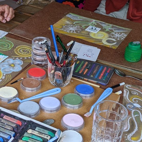 Ein Holztisch zeigt eine Vielzahl von Künstlerutensilien. Es gibt Pinseln in einem Glasbecher, verschiedene Farben in kleinen Behältern und ein Glas Wasser. Kunstpapiere mit Zeichnungen sind herum verstreut.