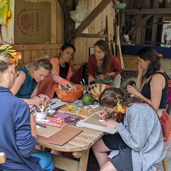 Eine Gruppe von Frauen sitzt um einen Holztisch herum und ist mit Kunstaktivitäten beschäftigt. Verschiedene Kunstutensilien, darunter Stifte, Bleistifte und eine Schüssel, sind auf dem Tisch verteilt. Eine Frau in der Mitte hält einen Stift und schreibt auf ein Blatt Papier.