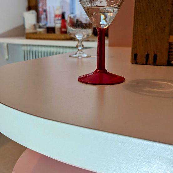 Zwei Weingläser stehen auf einer Theke. Ein Glas ist leer und das andere enthält Wasser. Hinter den Gläsern steht ein Holztablett mit Gegenständen darauf.