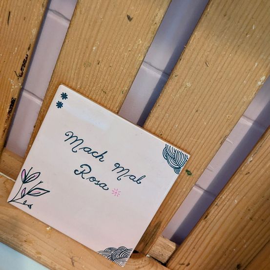 Ein quadratischer Keramikfliese mit einem Blumendesign auf einer Holzoberfläche. Die Fliese hat den Text 'Mach mal Rosa' in Blau.