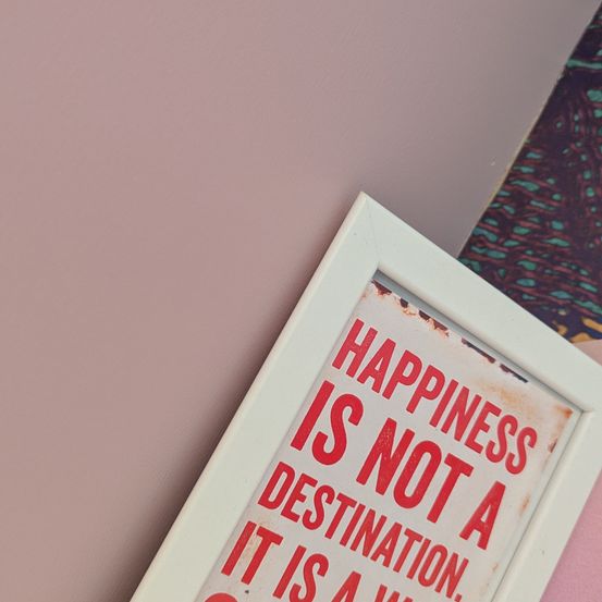 Ein gerahmter Spruch an einer rosa Wand sagt: 'GLÜCK IST KEIN ZIEL. ES IST EIN WEG.' Der Hintergrund ist ein Gradient von Rosa und Blaugrün.