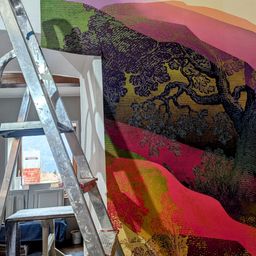 Eine Leiter lehnt an einer Wand mit einem farbenfrohen Landschaftswandbild. Das Wandbild zeigt einen Baum und Hügel in leuchtenden Farben. Links befindet sich ein Fenster mit einem Honda-Schild.