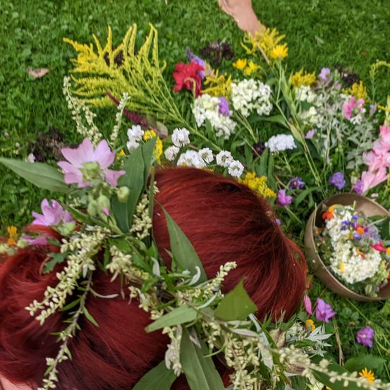 Eine Person mit roten Haaren, geschmückt mit einer Blumenkrone, liegt auf einem Grasfeld mit einer Vielzahl bunter Blumen darum. Ein kleiner Korb mit Blumen ist in der Nähe.