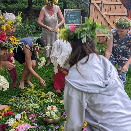 Eine Gruppe von Menschen ist auf einem Rasen und ordnet Blumen an. Einige tragen Blumenkränze. Eine Frau steht mit einem Blackboard in der Nähe.