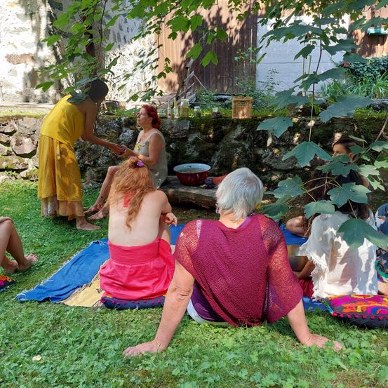 Eine Gruppe von Frauen sitzt im Kreis auf dem Gras und trägt bunte Kleidung. Eine Frau massiert die Füße einer anderen Frau. Hinter ihnen befindet sich eine Steinmauer und Pflanzen.