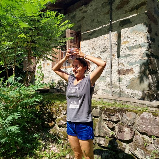 Eine Frau in einem grauen Tanktop und blauen Shorts steht draußen, hält eine Flasche und lacht vor einer Steinmauer und Pflanzen.
