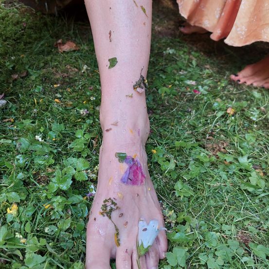 Ein Nahaufnahme eines nackten Fußes, der mit Blättern und Blumen auf einem Grasfeld bedeckt ist.