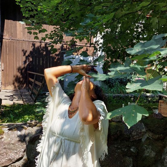 Eine Frau in Weiß steht unter einem Baum und schaut nach oben. Sie scheint Blätter zu pflücken. Dahinter ist ein Holzzaun zu sehen, und links befindet sich eine Holzstruktur.