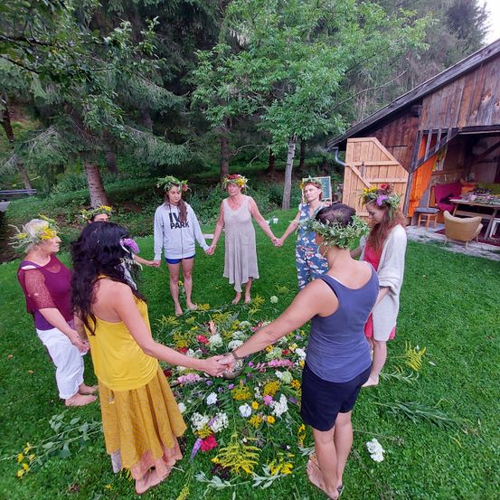 Eine Gruppe von Frauen mit Blumenkränzen hält sich an den Händen in einem Kreis um eine Blumendekoration auf einer Rasenfläche. Hinter ihnen ist ein Holzhaus und Bäume zu sehen.