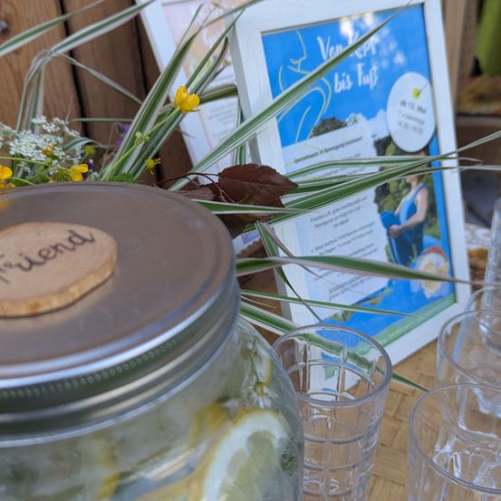 Ein Glas mit Zitronenscheiben und einem Korkdeckel, beschriftet mit 'friend', steht neben einem Poster und Gläsern auf einem Holztisch.