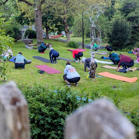 Eine Gruppe von Menschen praktiziert Yoga auf einem Rasen, umgeben von Bäumen und Pflanzen. Einige knien, andere stehen, alle konzentrieren sich auf ihre Übung. Yogamatten sind auf dem Boden ausgelegt.