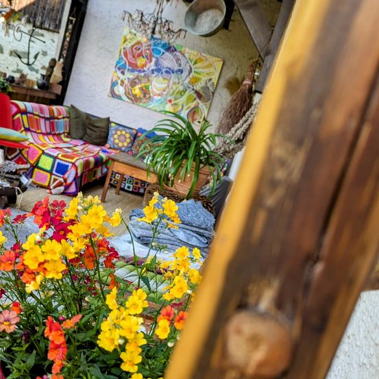 Ein gemütlicher Innenbereich mit einem bunten Sofa, einer Topfpflanze und einem abstrakten Gemälde an der Wand. Helle Blumen im Vordergrund.