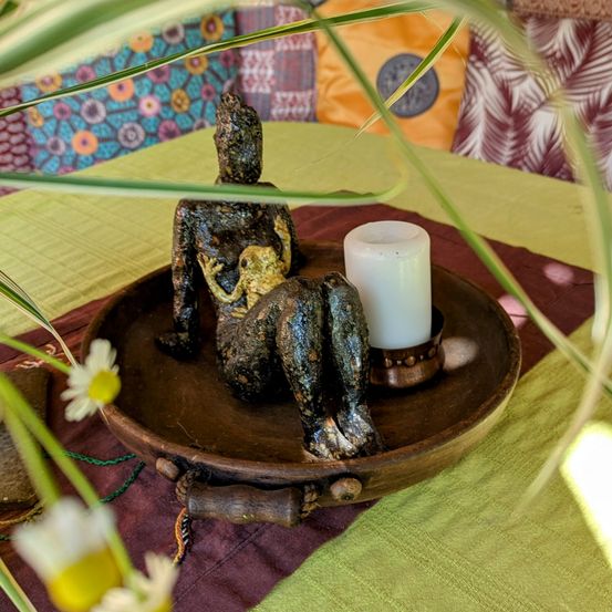 Ein hölzerner Tablett mit einer kleinen Statue einer sitzenden Frau, die einen Frosch auf ihrem Schoß hält, eine Kerze daneben und umgeben von Pflanzen.