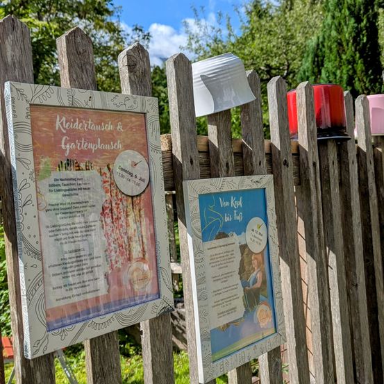 Ein Holzzaun mit zwei gerahmten Postern. Ein Poster bewirbt einen Kleidungs- und Gartentausch für Sonntag, 6. Juli. Das andere Poster bewirbt eine Kessel-zum-Badewanne-Veranstaltung.