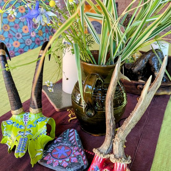 Ein Tisch mit einem dunklen Tischläufer hat einen dekorierten Schädel, Geweihe, einen Stein und eine grüne Vase mit einer Pflanze. Dahinter eine gemusterte Wand und eine weiße Kerze.