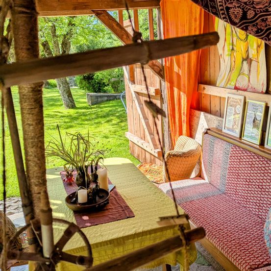 Eine rustikale Holzbank und ein Tisch mit einem gelben Tuch in einem Außenbereich. Dahinter eine Holzkonstruktion, mit einem Baum und einem Rasen.