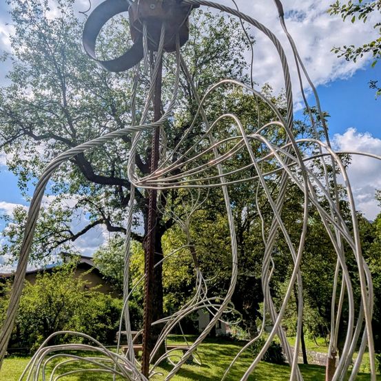 Eine Metallskulptur, die einer verdrehten Schleife ähnelt, steht in einem Park. Sie ist von üppigen Bäumen umgeben, unter einem blauen Himmel mit Wolken.
