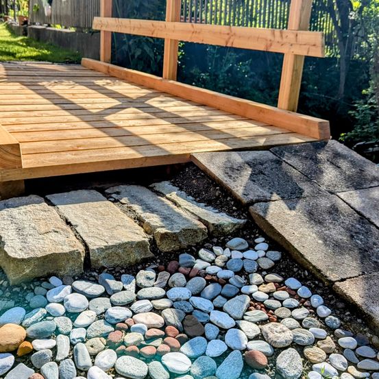 Eine Holzbrücke führt über einen kleinen Steinweg, mit Kieselsteinen und Felsen auf beiden Seiten. Die Brücke wird von Holzbalken gestützt, und Sonnenlicht filtert durch das umgebende Grün.