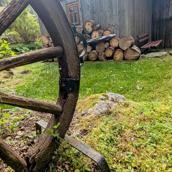 Ein hölzernes Rad mit einem rostigen Metallteil steht vor einem Haus. Das Haus hat ein Fenster und eine Tür. Vor dem Haus befindet sich ein Haufen Holzscheite und eine Holzbank.