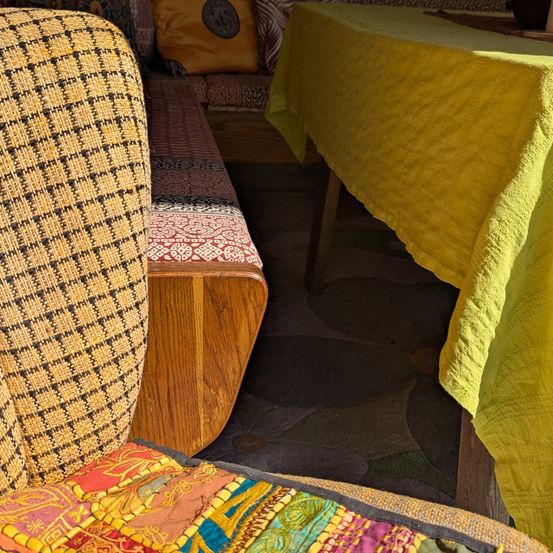 Ein Nahaufnahme von einem hölzernen Stuhl mit farbenfrohen gemusterten Kissen. Ein gelber Tischläufer bedeckt einen Tisch rechts, während ein beiges Kissen auf einer gepolsterten Bank hinter dem Stuhl ruht.