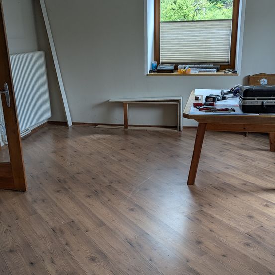 Ein leerer Raum mit Holzböden, einem Fenster mit Jalousien, einer Bank, einem Holztisch mit Werkzeugen und einer Reisetasche. An der Wand steht ein weißer Heizkörper.