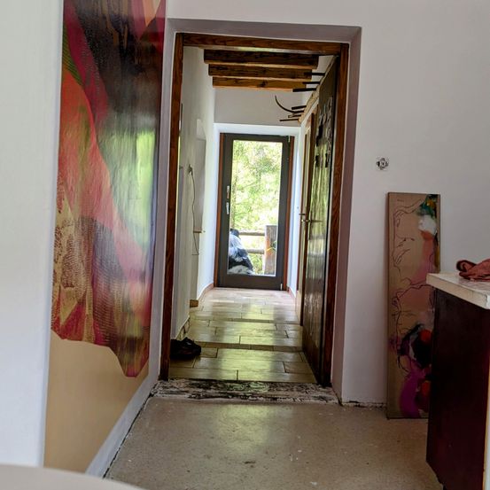 Ein Flur mit einem großen abstrakten Gemälde auf der linken Seite und einer offenen Tür, die zu einem Raum führt, in dem eine Person vor einem Fenster steht. Ein Teppich liegt am Eingang.