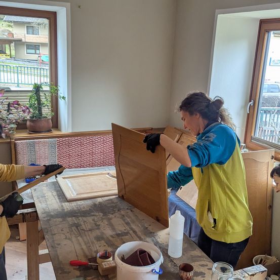 Zwei Personen arbeiten in einem Raum mit Fenster und Bank an einem Holzschrank. Eine Person hält eine Holzplatte, während die andere am Schrank arbeitet. Auf der Fensterbank steht eine Pflanze und draußen steht ein Auto.
