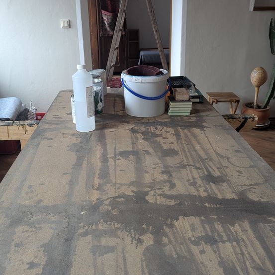 Ein Arbeitsbereich mit einem langen Tisch, der Spuren von Malerei zeigt. Ein weißer Eimer und Farbdosen sind auf dem Tisch, zusammen mit einigen Büchern. Eine Leiter und ein Topf mit Pflanzen im Hintergrund.