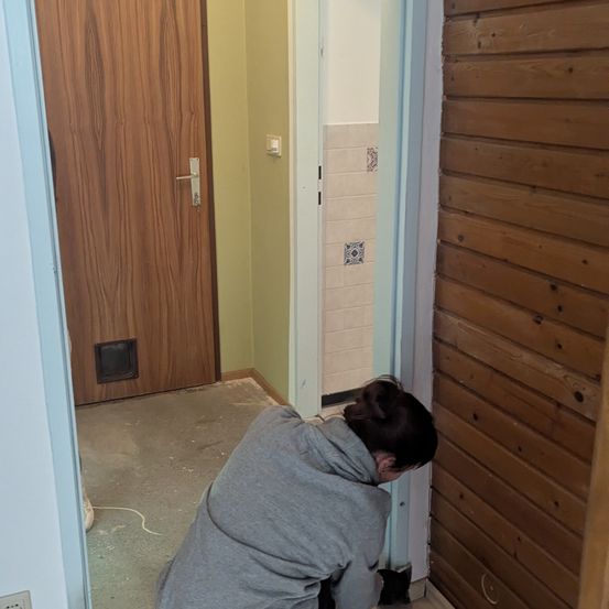 Eine Frau kniet auf dem Boden und scheint etwas zu reparieren. Sie befindet sich in einem Raum mit einer Holztür und gefliesten Wänden.