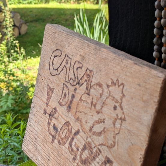 Ein Holzblock mit 'Casa Di Gocce' und einem eingravierten Hahn steht im Garten. Dahinter befinden sich Pflanzen und Holzscheite.