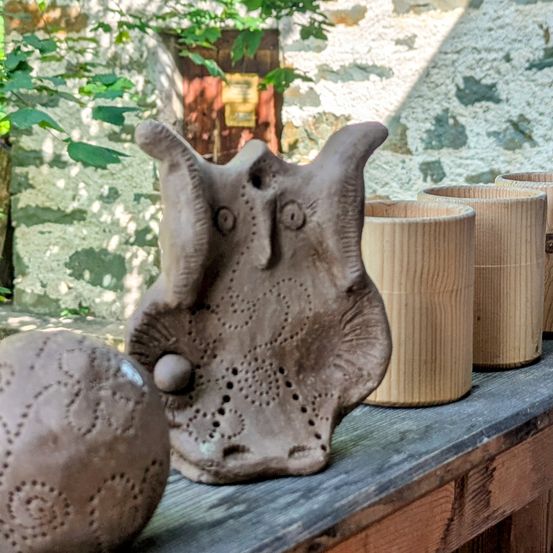 Eine Tonfigur einer Eule steht auf einem Holztisch mit vier Holzbechern, vor einer Steinmauer und Grünpflanzen.