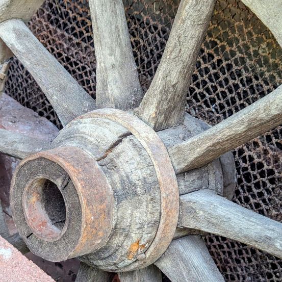 Ein Nahaufnahme eines alten hölzernen Wagenrads zeigt verrostetes Metall um die Achse, mit hölzernen Speichen und einem verwitterten Rand.