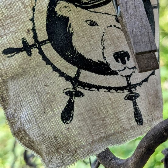 Ein Jutetuch mit einer Bärenillustration hängt an einer Holzklammer. Der Bär ist mit Hut und einem Lenkrad dargestellt.