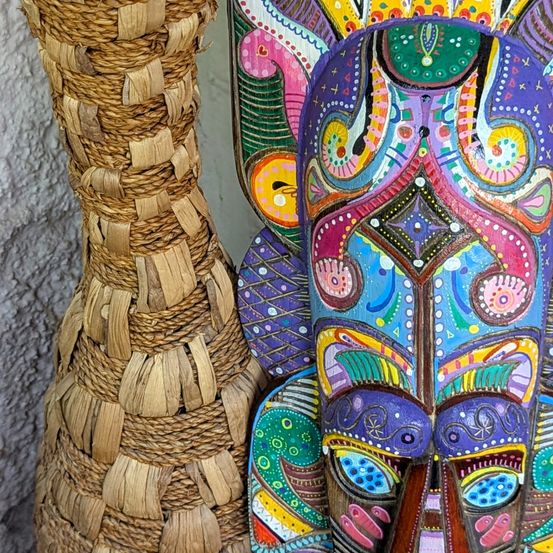 Eine farbenfrohe, kunstvoll gestaltete Holzmaske mit komplexen Mustern steht neben einem gewebten, braunen Bambusvase. Die Maske zeigt kräftige, abstrakte Designs in Lila, Grün, Gelb und Blau.