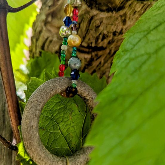 Ein holzner Ring mit bunten Perlen hängt von einem Ast, eingebettet zwischen üppigen grünen Blättern.