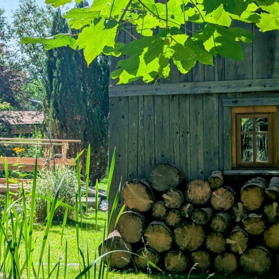 Ein rustikales Holzhaus mit einem kleinen Fenster und einem Stapel Brennholz ist von üppiger Grünfläche und Bäumen umgeben. Im Hintergrund befindet sich eine Holzstruktur mit einer gelben Blume.