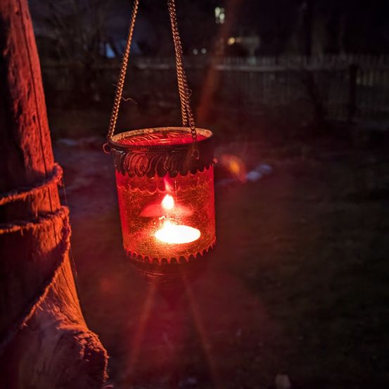 Ein Laterne mit rotem Glas und einer brennenden Kerze hängt an einem Baumzweig in einem dunklen Bereich.
