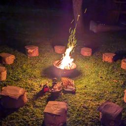 Ein Lagerfeuer brennt hell in einem Kreis aus Baumstämmen auf einem Grasfeld, umgeben von Pappkartons. Es ist Nacht.
