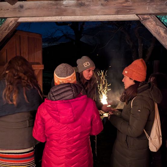 Fünf Frauen versammeln sich bei Nacht unter einem Holzpavillon, eine Frau hält eine Wunderkerze, während andere zusehen.