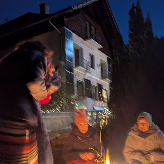 Drei Menschen sitzen bei einem Feuer vor einem Haus in der Nacht. Eine Frau lehnt sich über das Feuer, während die anderen beiden zurücksitzen. Das Haus hat einen Schornstein, Fenster und einen Balkon.