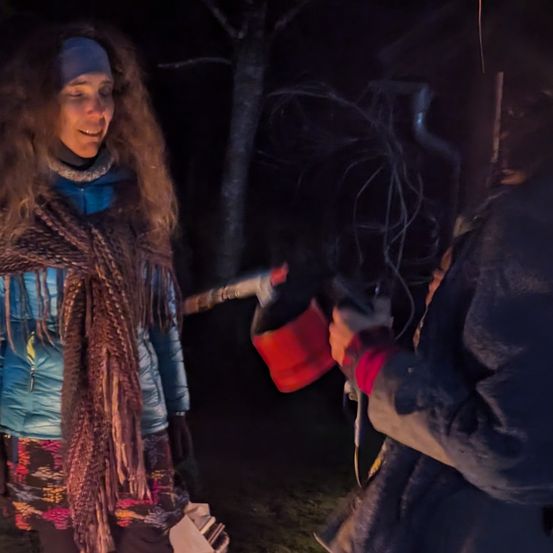 Zwei Personen stehen bei einem Lagerfeuer in der Nacht. Eine Person hält ein rotes Objekt, möglicherweise eine Fackel. Sie sind beide warm gekleidet.