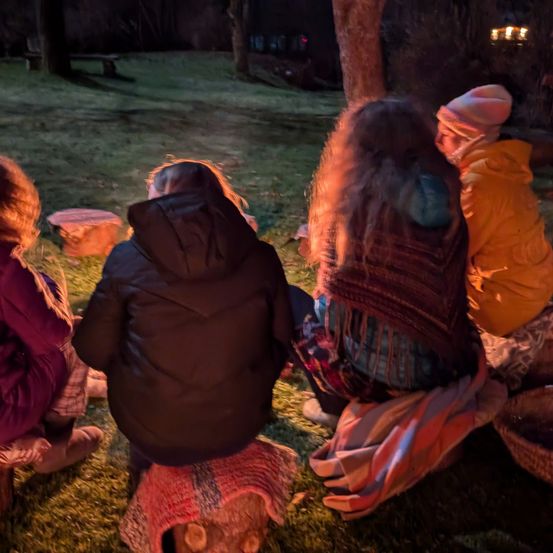 Vier Menschen sitzen im Kreis auf dem Gras in der Nähe eines Lagerfeuers, gekleidet in warme Kleidung. Zwei von ihnen tragen Mützen, und einer hat eine Schürze. Der Hintergrund ist schwach beleuchtet mit einer Bank und entfernten Lichtern.