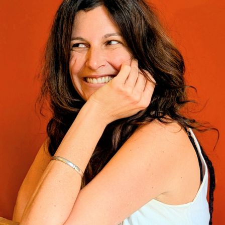 Eine Frau mit langen, welligen Haaren lächelt und posiert mit der Hand am Kinn vor einem roten Hintergrund. Sie trägt ein weißes ärmelloses Top und eine silberne Armband.